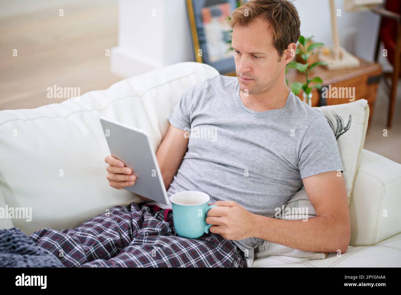 Ein entspannter Tag zu Hause. Ein gutaussehender Mann, der Kaffee trinkt, während er sein Tablet zu Hause verwendet. Stockfoto