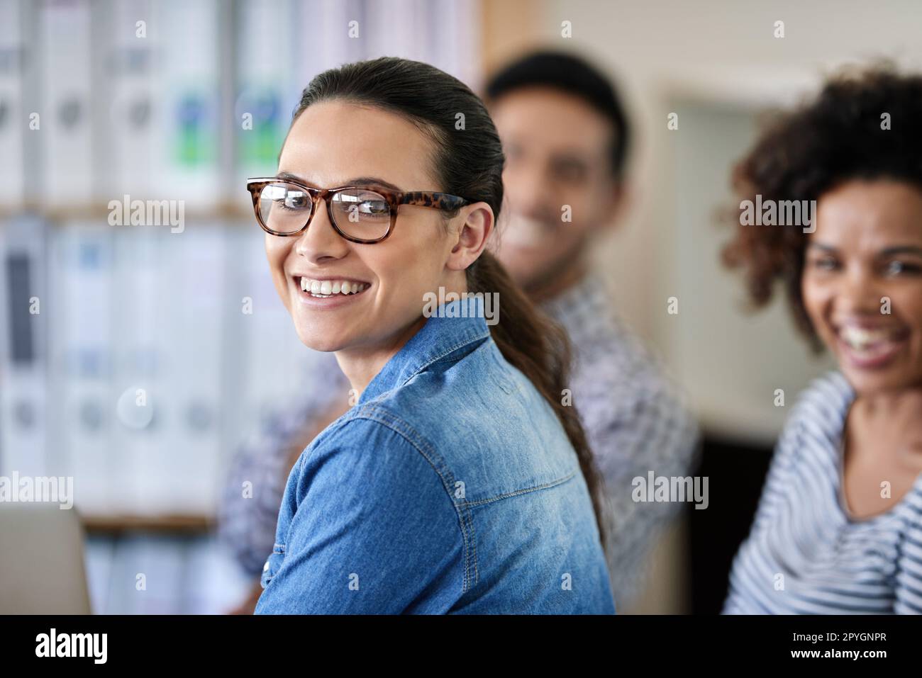 Das Arbeitsleben ist keine lästige Pflicht. Eine Geschäftsfrau lächelt in der Kamera mit ihren Kollegen im Hintergrund. Stockfoto