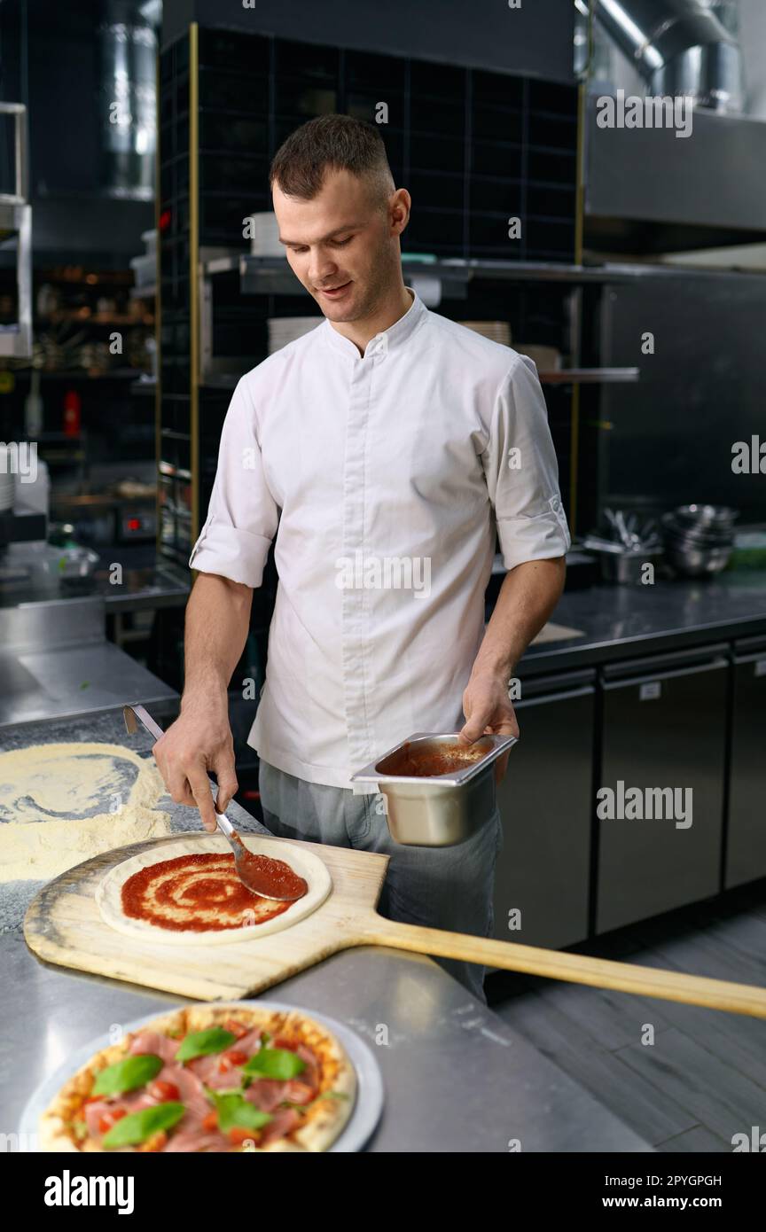 Männlicher Koch, der Tomatenpaste auf Pizzabasis ausstreut Stockfoto