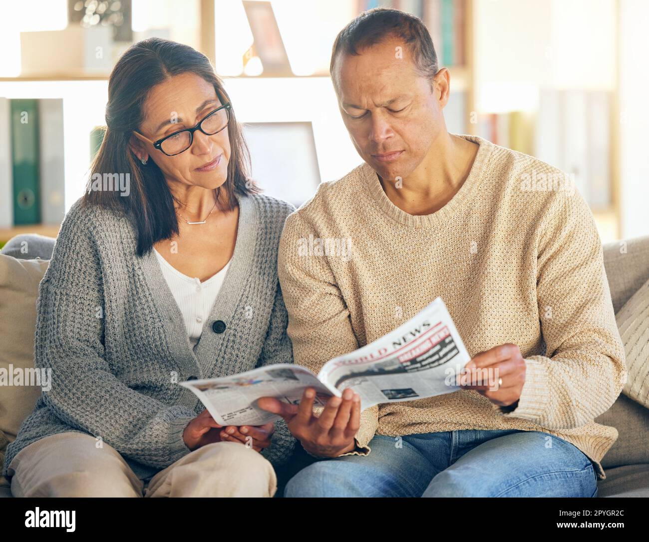 Seniorenpaar, Lesen und Zeitung auf dem Sofa, konzentrieren oder entspannen Sie sich zusammen am Morgen für eine globale Veranstaltung. Reife Männer, Frauen oder Medien für internationale Nachrichten, Informationen oder Weltkrisen auf der Couch Stockfoto