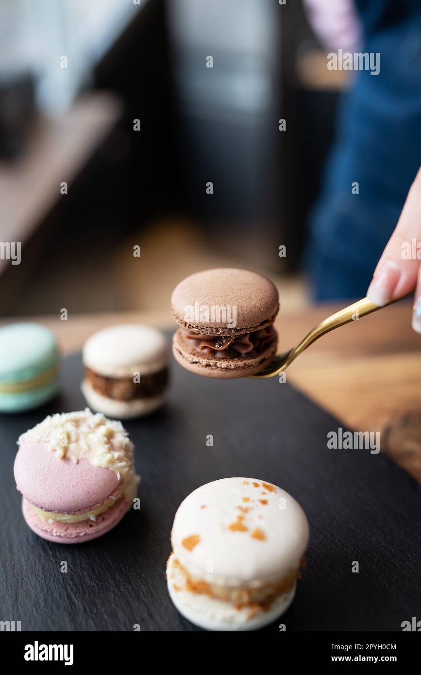 Ein köstliches Makkaron-Dessert - eine süße französische Delikatesse liegt auf einem schwarzen Betonständer, und ein Mädchen hält ein Bonbon mit einer Gabel. Vertikales Foto. Stockfoto