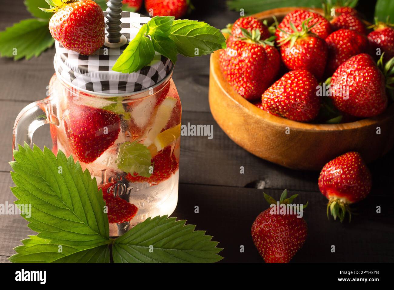 Sommerliches kaltes Getränk mit Minze, Eis, Zitrone und Erdbeeren und einer Holzschale mit frischen Erdbeeren auf schwarzer Holzoberfläche. Stockfoto