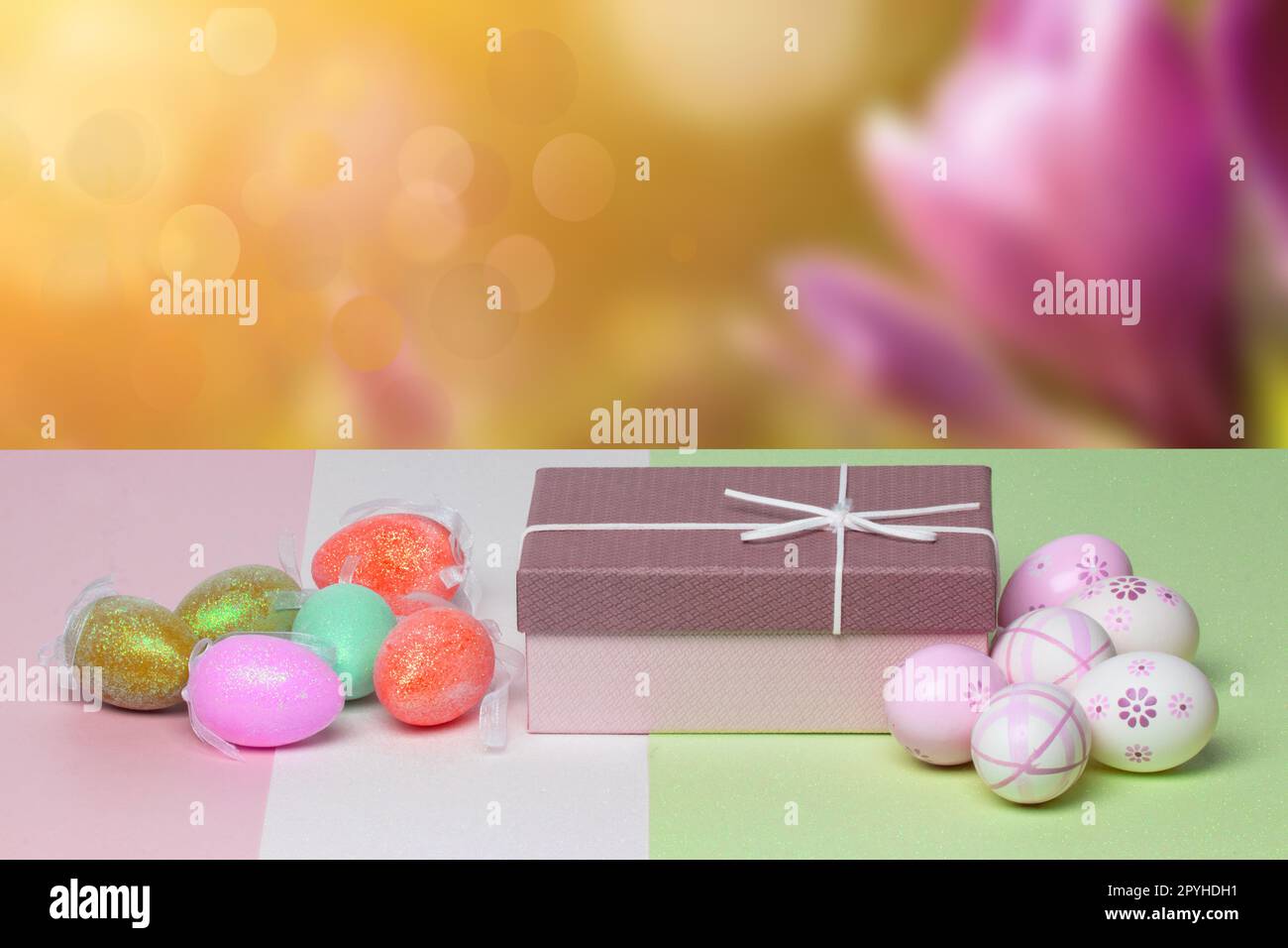 Vorlage für Ostergrußkarte. Nahaufnahme von bunten Eiern und einer Geschenkbox auf einem mehrfarbigen Tisch vor abstraktem Frühlingshintergrund. Kopierbereich für Ihr Textdesign. Stockfoto