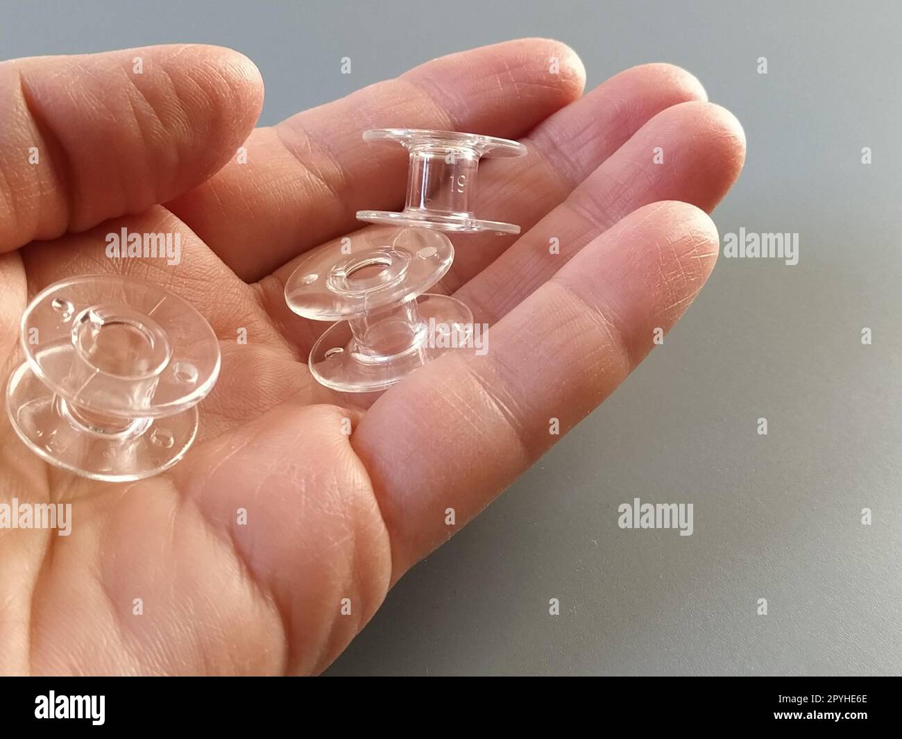 Transparente Kunststoffspulen einer modernen Nähmaschine in der Handfläche einer Frau - eine Näherin. Grauer Hintergrund. Nähmaschinen Stockfoto