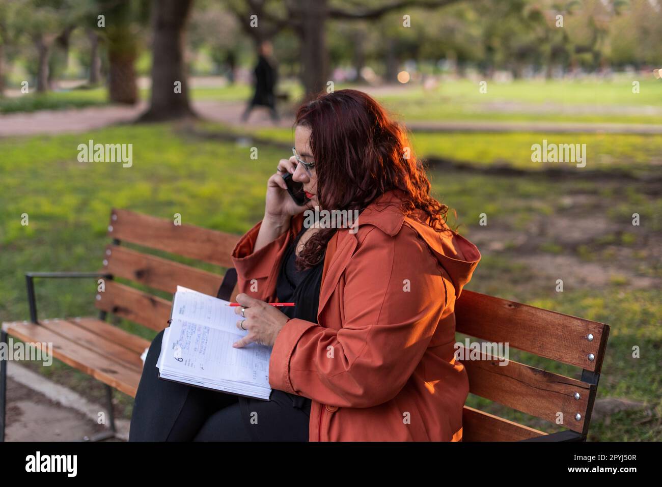 Eine Executive Plus-Size-Frau, die im Freien arbeitet. Sie schreibt auf ihr Notebook und führt ein Telefongespräch, während sie in der Öffentlichkeit auf einer Bank sitzt Stockfoto