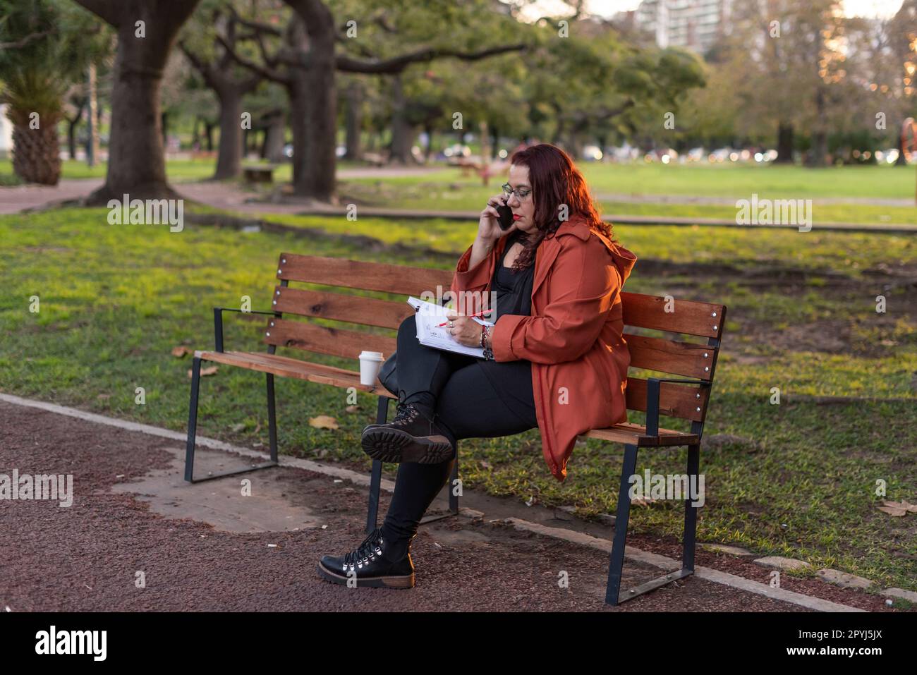 Eine Executive Plus-Size-Frau, die im Freien arbeitet. Sie schreibt auf ihr Notebook und führt ein Telefongespräch, während sie in der Öffentlichkeit auf einer Bank sitzt Stockfoto