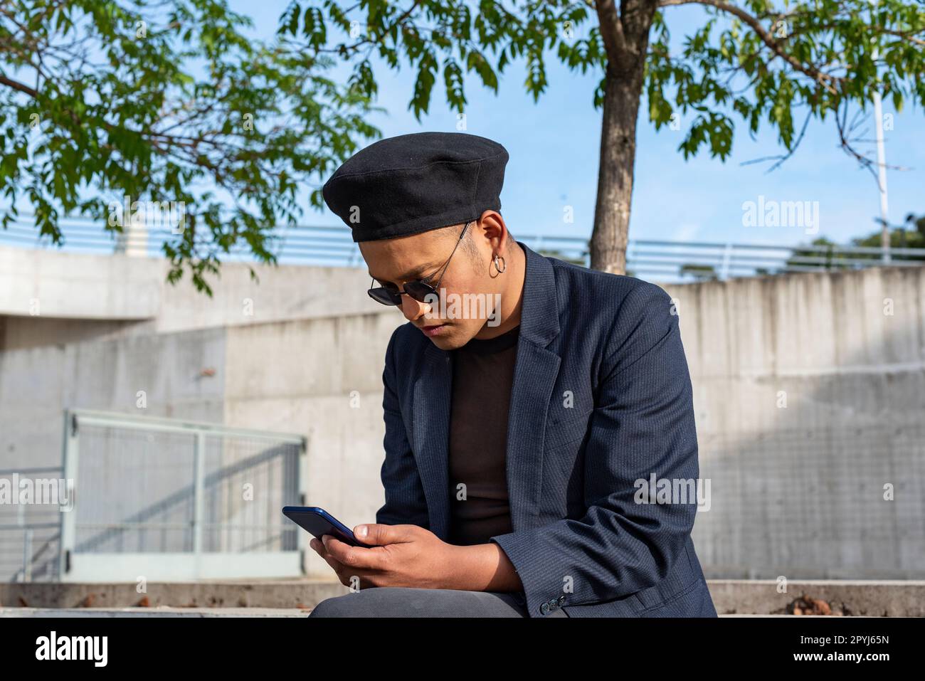 Ein junger lateinischer Schwuler mit modischem Hut und Sonnenbrille, der im Freien am Strand SMS auf seinem Smartphone schreibt. LGTB. Stockfoto