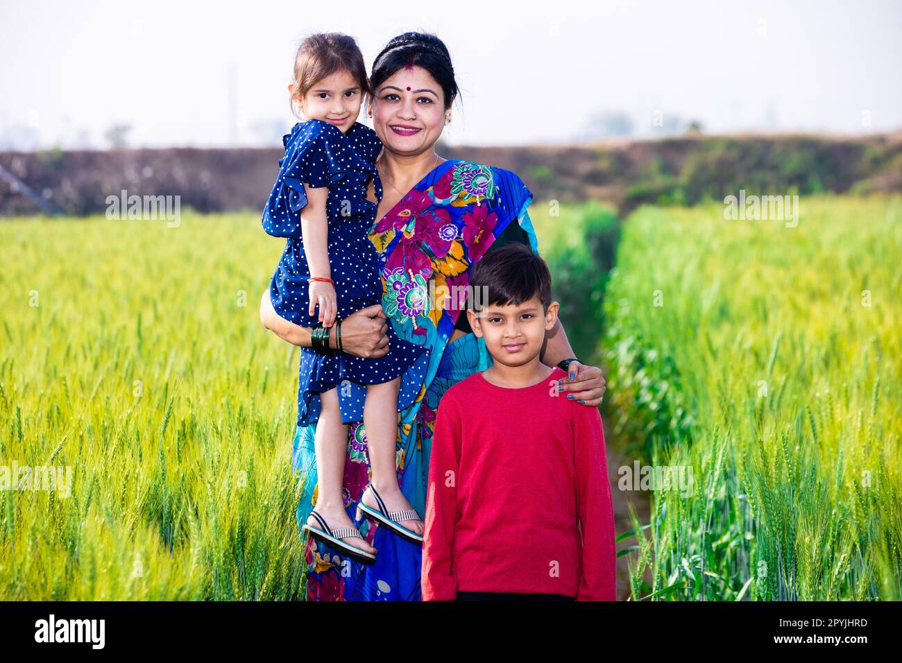 Glückliche junge, traditionelle indische Mutter, die mit ihren Kindern auf Weizenlandwirtschaft steht, Konzept für das ländliche indien. Stockfoto