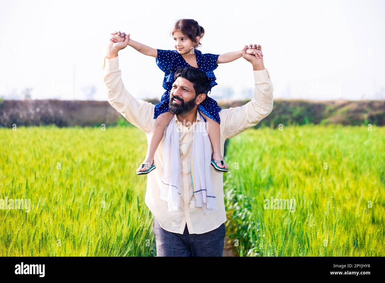 Glückliche kleine indische Tochter, die auf der Schulter des Vaters sitzt, Spaß beim Gehen auf Weizenlandwirtschaft hat, Freizeit zusammen auf der Farm verbringt, Mädchen Kind Stockfoto