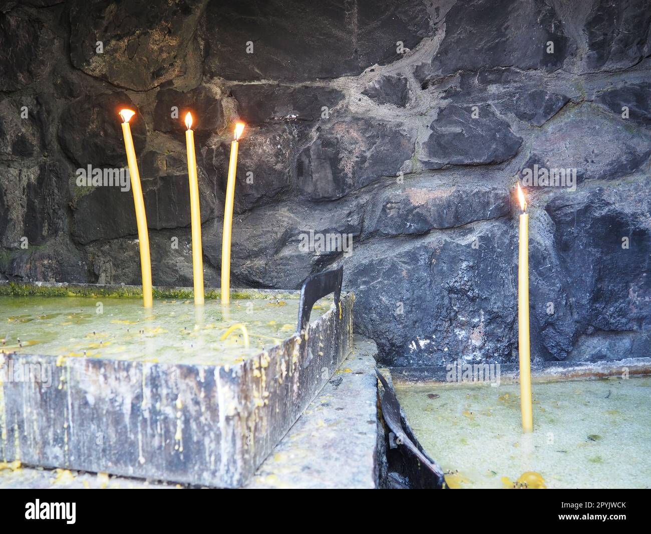 Brennende Kerzen für Frieden oder Gesundheit. Religiöser Ritus der Kirche. Drei Kerzen auf der linken Seite, eine auf der rechten Seite. Geschmolzenes Wachs in einem Metallbehälter. Steinhintergrund Stockfoto