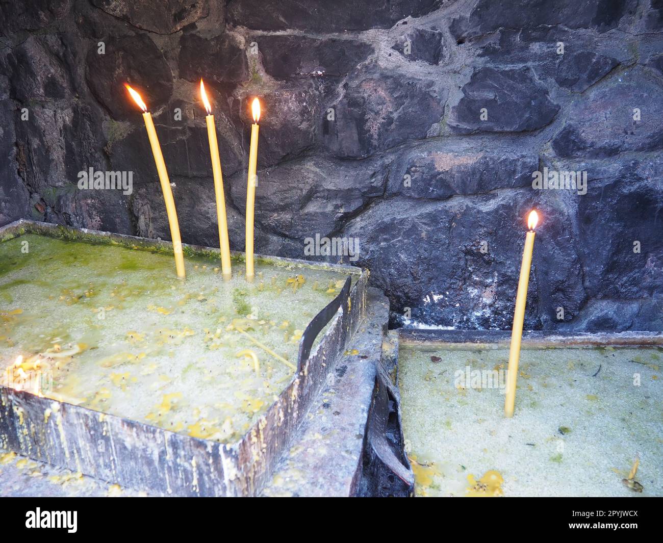 Brennende Kerzen für Frieden oder Gesundheit. Religiöser Ritus der Kirche. Drei Kerzen auf der linken Seite, eine auf der rechten Seite. Geschmolzenes Wachs in einem Metallbehälter. Steinhintergrund. Stockfoto