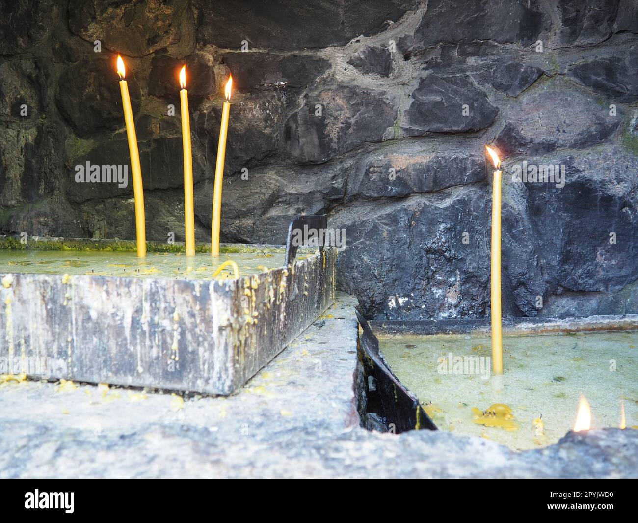 Brennende Kerzen für Frieden oder Gesundheit. Religiöser Ritus der Kirche. Drei Kerzen auf der linken Seite, eine auf der rechten Seite. Geschmolzenes Wachs in einem Metallbehälter. Steinhintergrund. Stockfoto