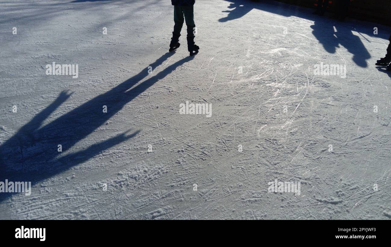 Kinder fahren in einem Stadtpark auf einer Eisbahn. Beim Eislaufen laufen laufen laufen die Füße. Die niedrige Wintersonne erhellt das Eis schwach. Dunkle Formen und lange Schatten auf der Oberfläche. Sportbewegungen Stockfoto