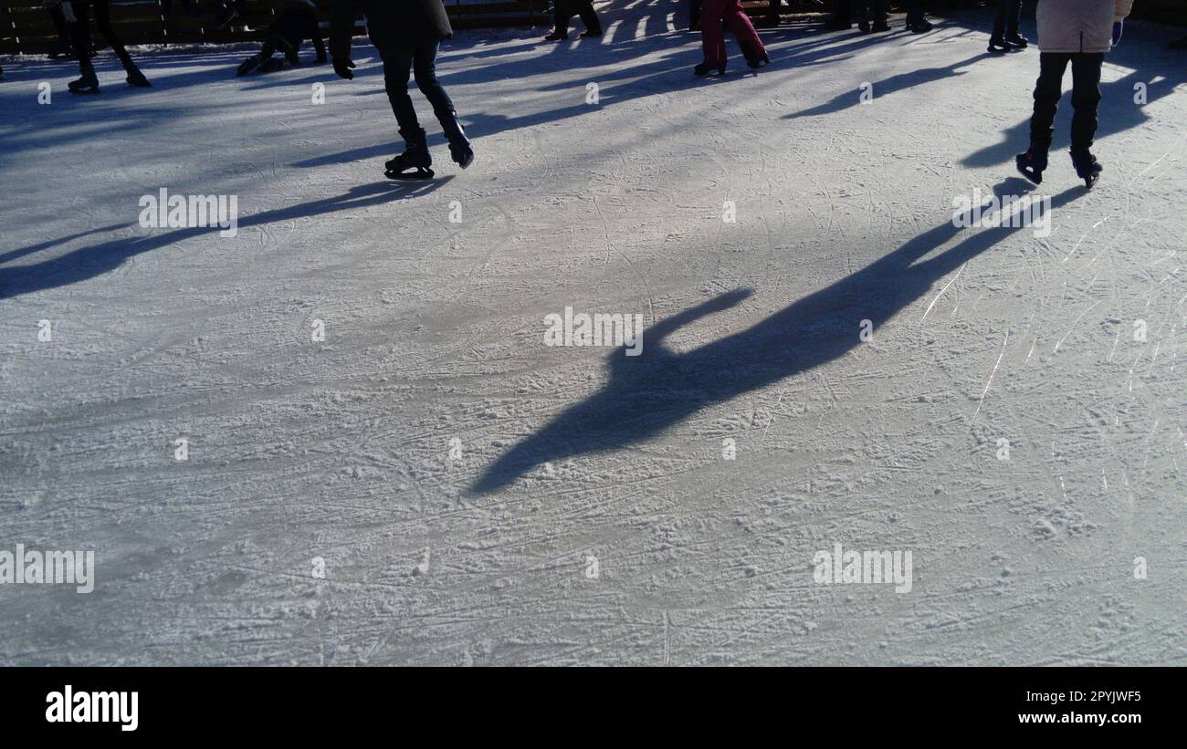 Kinder fahren in einem Stadtpark auf einer Eisbahn. Beim Eislaufen laufen laufen laufen die Füße. Die niedrige Wintersonne erhellt das Eis schwach. Dunkle Formen und lange Schatten auf der Oberfläche. Sportbewegungen Stockfoto