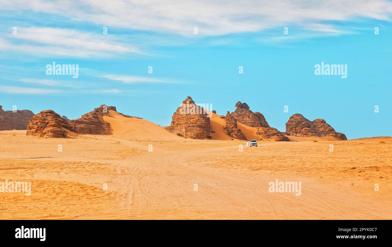 Typische Wüstenlandschaft in Alula, Saudi-Arabien, Sand mit einigen Bergen, kleines Geländefahrzeug, einheimischer Mann und Kamele in der Ferne Stockfoto