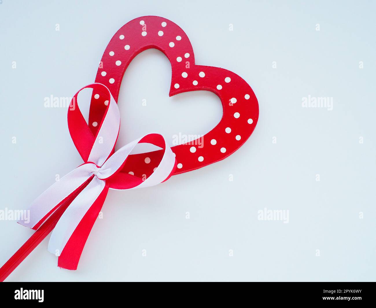 Valentinstag-Konzept. Rotes Holzherz mit weißen Punkten und Schleife an einem Stock. Weißer Hintergrund. Eine schöne Schleife als Dekoration. Liebe und Beziehungen. Anerkennung oder Glückwünsche Stockfoto