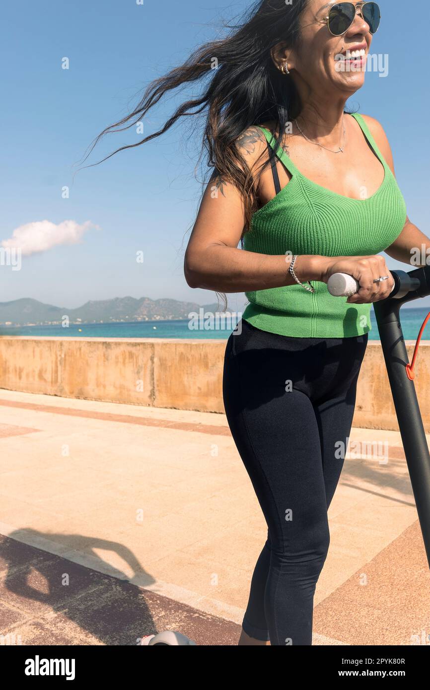 Junge Latina-Frau, die Spaß mit dem elektrischen Roller am Strand hat Stockfoto