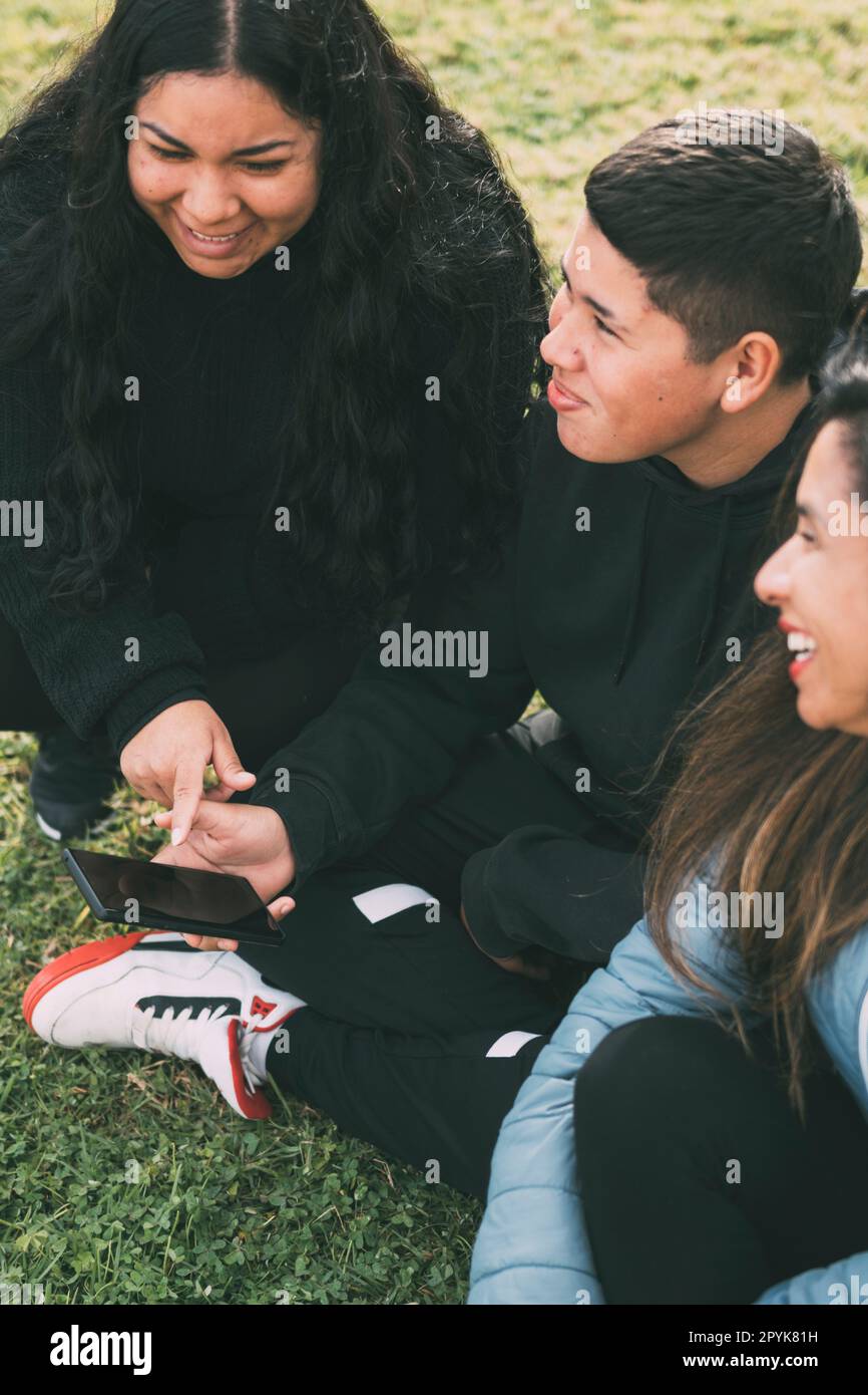 Drei Menschen hispanisch-lateinamerikanischer Herkunft, die mit intelligenten Geräten auf dem Boden des Parks sitzen Stockfoto