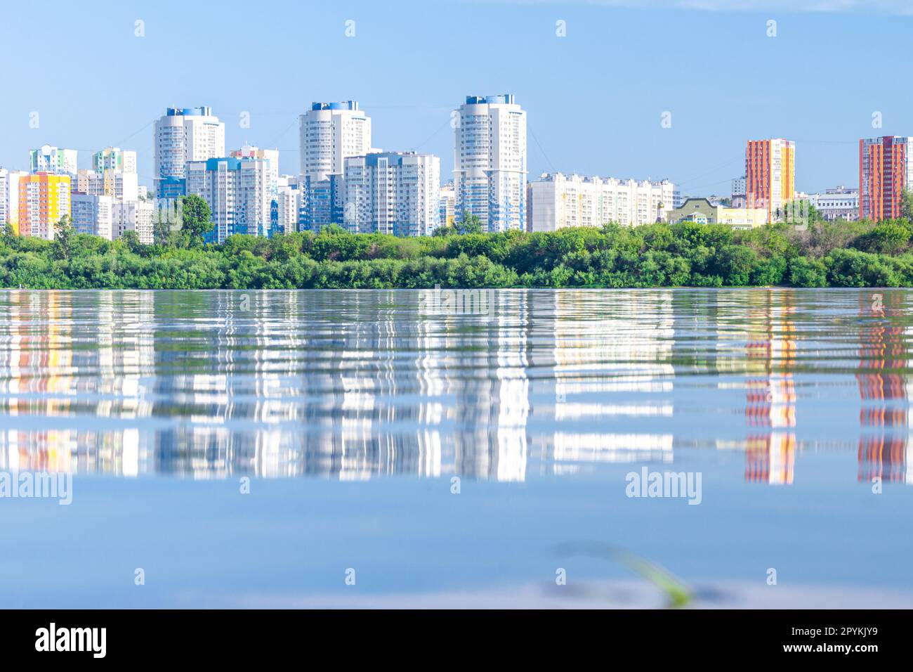 Rot-gelbe und blau-weiße Hochhäuser hinter der Küstenvegetation spiegeln sich im Wasser eines Sees oder Flusses wider, selektiver Fokus Stockfoto