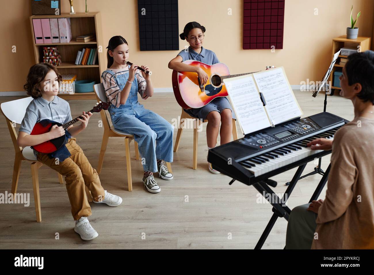 Gruppe von Kindern, die auf Stühlen sitzen und Musikinstrumente spielen, während der Lehrer während des Unterrichts Klavier spielt Stockfoto