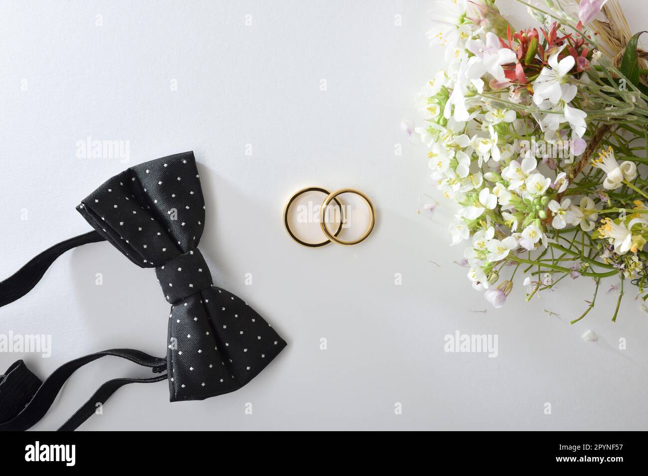 Hochzeitskonzept mit goldenen Ringen in der Mitte auf weißem Tisch und schwarzer Fliege und Blumenstrauß. Draufsicht. Stockfoto