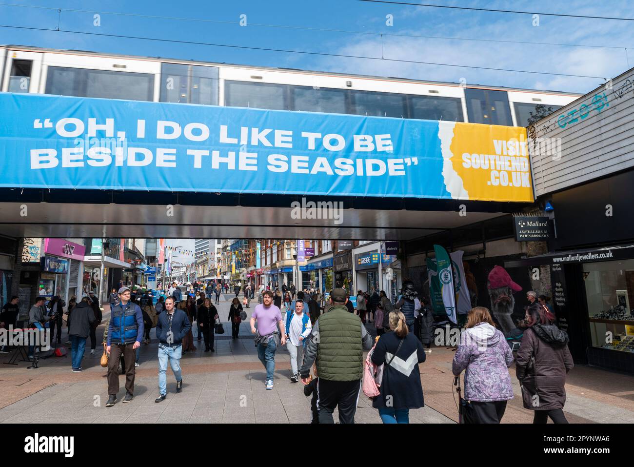 Leute in der High Street, Southend auf dem Meer, die unter der Eisenbahnbrücke vorbeifahren mit "Oh, ich mag es, am Meer zu sein". Zug um C2C Uhr auf der Brücke. Beschäftigt Stockfoto