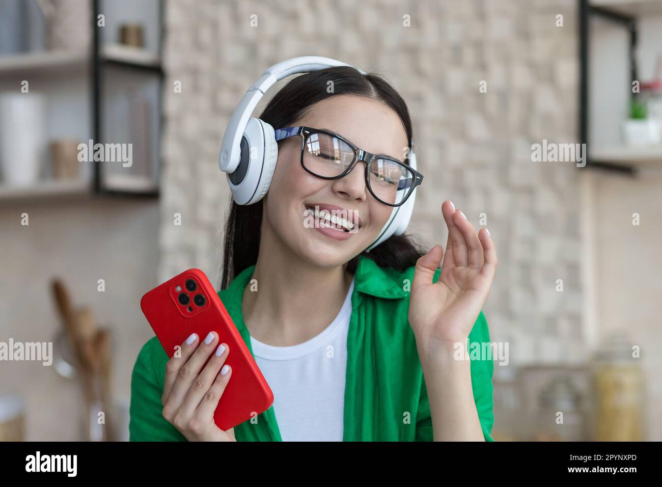 Nahaufnahme. Eine lächelnde junge Studentin hört zu Hause Musik über Kopfhörer und hält ein Telefon in der Hand. Sie schloss die Augen, ruht sich aus, genießt es, tanzt. Stockfoto