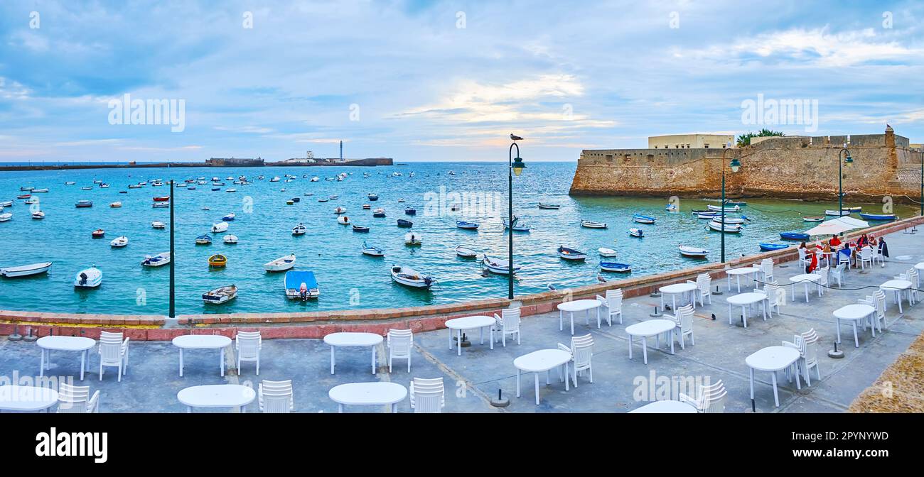 CADIZ, SPANIEN - 21. SEPTEMBER 2019: Panoramablick auf die Promenade La Caleta mit Speisen im Freien, vielen festgefahrenen Booten und mittelalterlichen Burgen im Hintergrund, am September Stockfoto