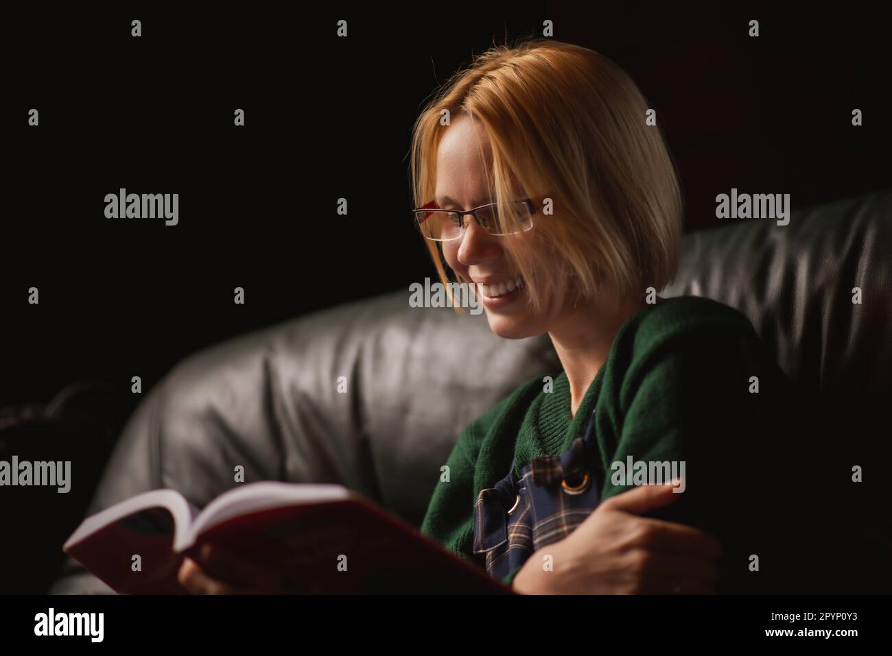 Eine Frau mit Brille lächelt und liest zu Hause ein Buch auf dem Sofa. Vierzig Jahre alte Frau liest ein faszinierendes Buch. Stockfoto