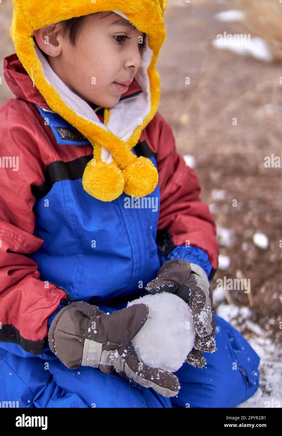Ein nachdenklicher lateinischer kleiner Junge kniet im Schnee mit einem Schneeball in den Händen, trägt einen Schneeanzug, einen gelben Hut und Handschuhe. Stockfoto
