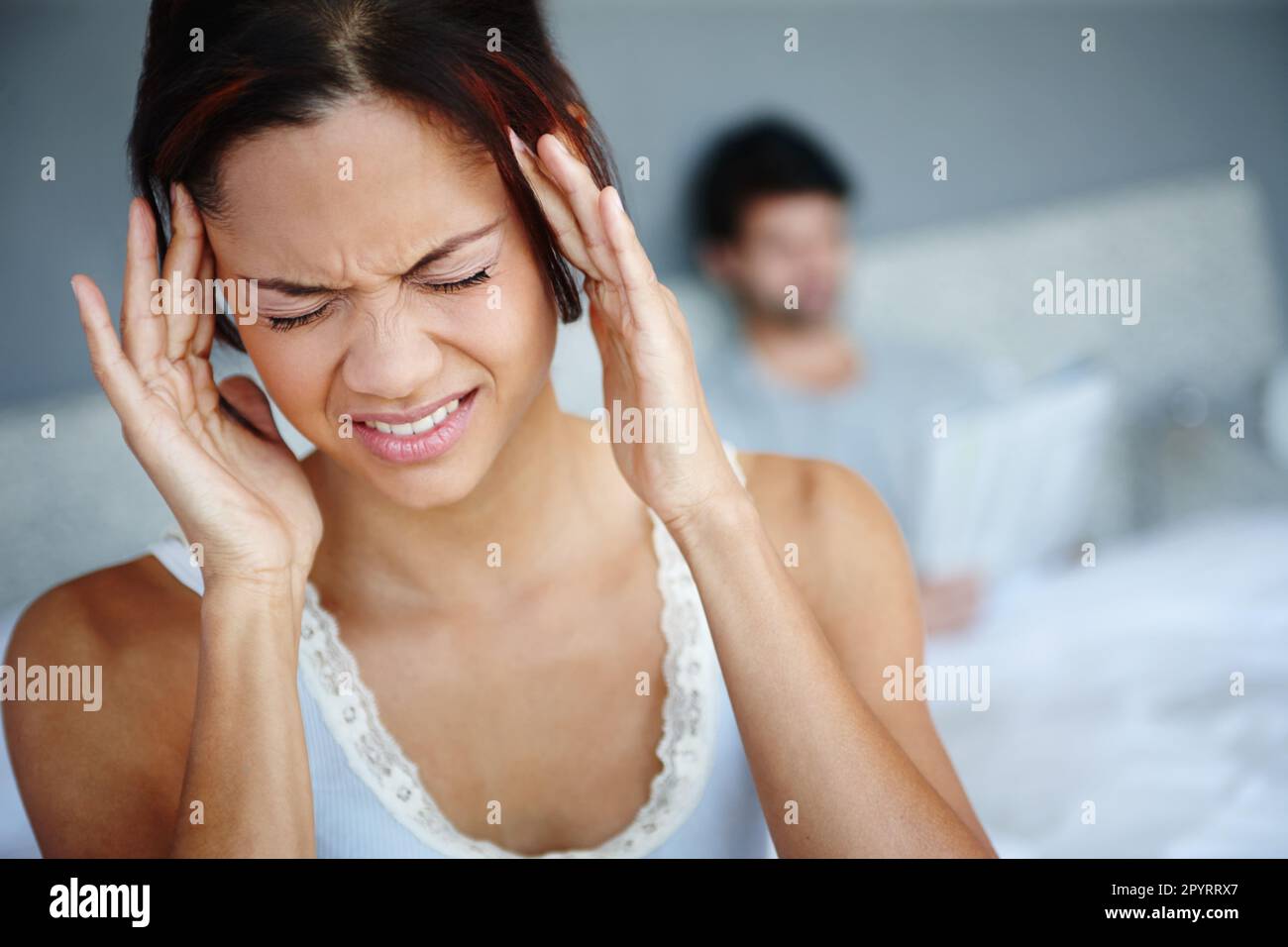 Migräneschmerzen. Eine Frau mit schlimmen Kopfschmerzen, die auf ihrem Bett sitzt, mit ihrem Freund im Hintergrund. Stockfoto
