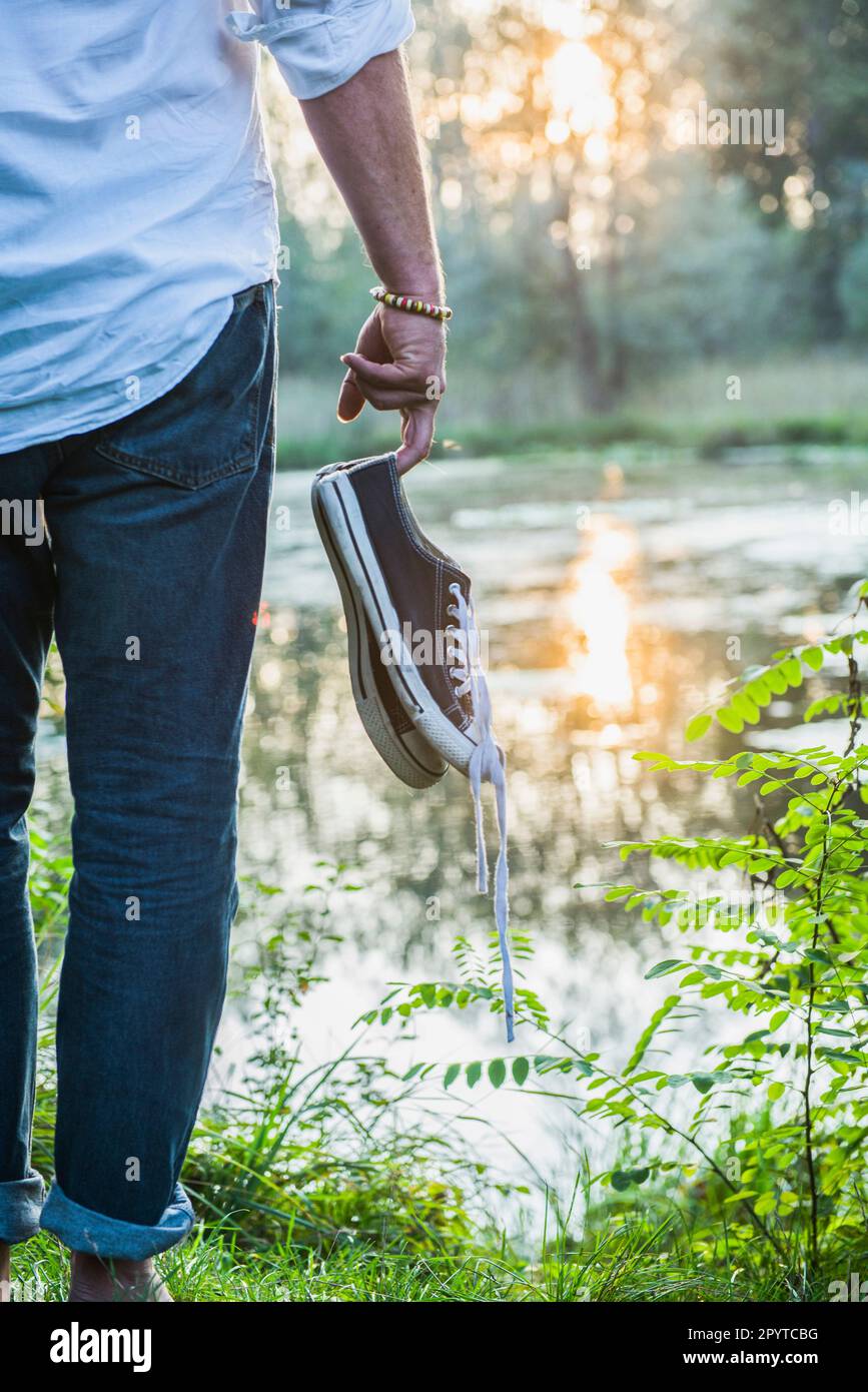 Mittelteil eines Mannes, der mit Schuhen in der Hand am Seeufer in Bayern steht Stockfoto