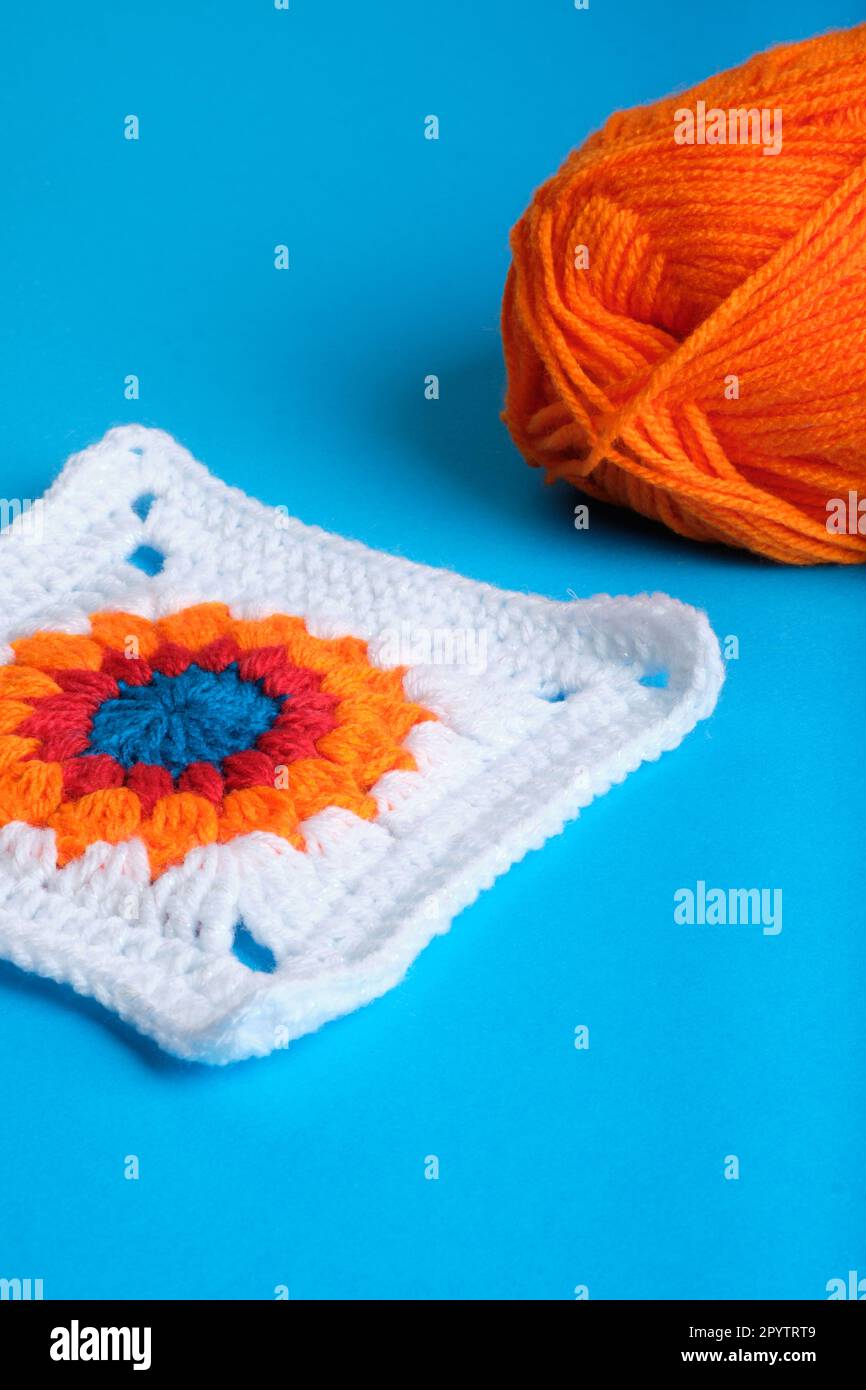 Granny Square mit Häkeldesign und weißem Quadrat mit orangefarbenem, rotem und blauem Muster in der Mitte und orangefarbenem Garnknäuel auf blauem Hintergrund. Stockfoto