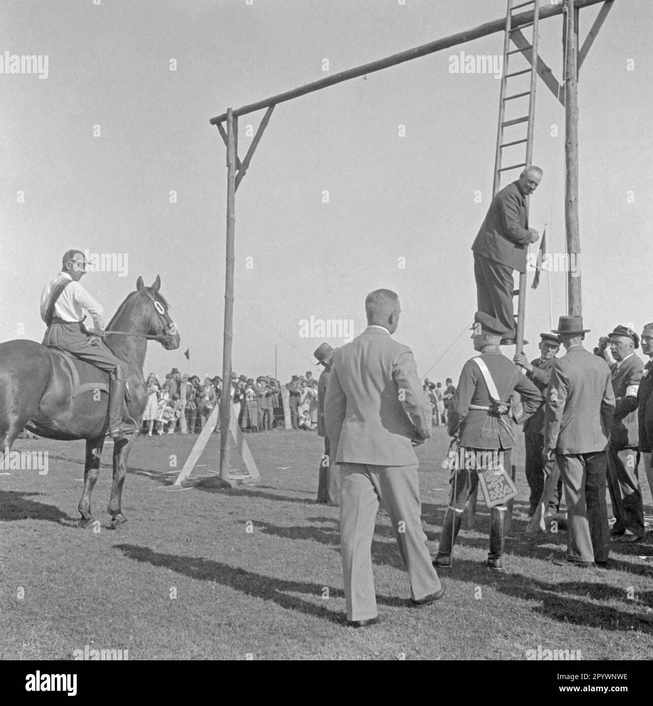 Ringreiten in Warnemünde. Beim Ringfahren wird eine Lanze durch einen Ring gesteckt, der an einem Gerüst hängt. Unter den Zuschauern ist ein Mann in preußischer Uniform. Stockfoto