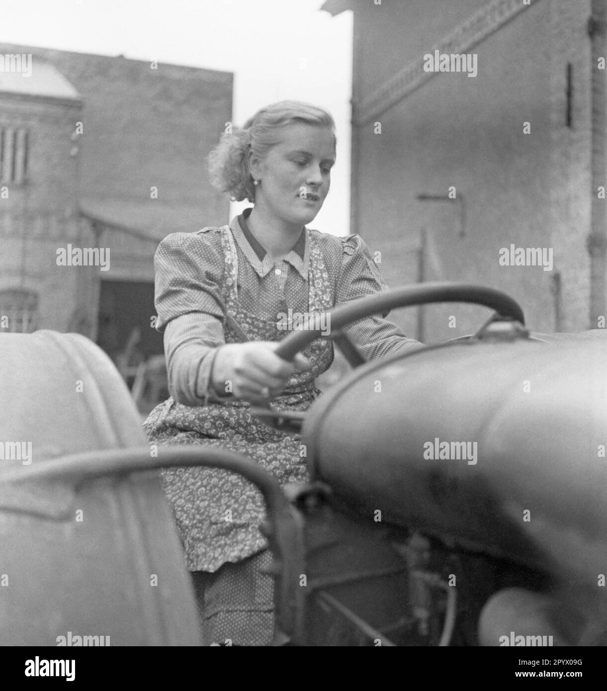 Eine blonde Frau, die ein Kleid und eine Schürze trägt, fährt einen Traktor. Im Hintergrund: Innenhofgebäude. Unbefristetes Foto. Stockfoto