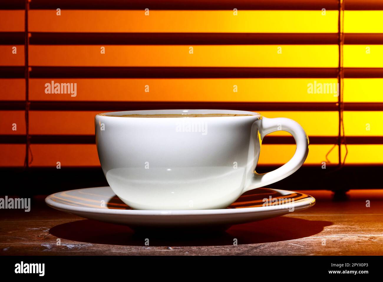 Kaffeetasse gefüllt mit schaumigem Kaffee, an einem Fensterrollo Stockfoto