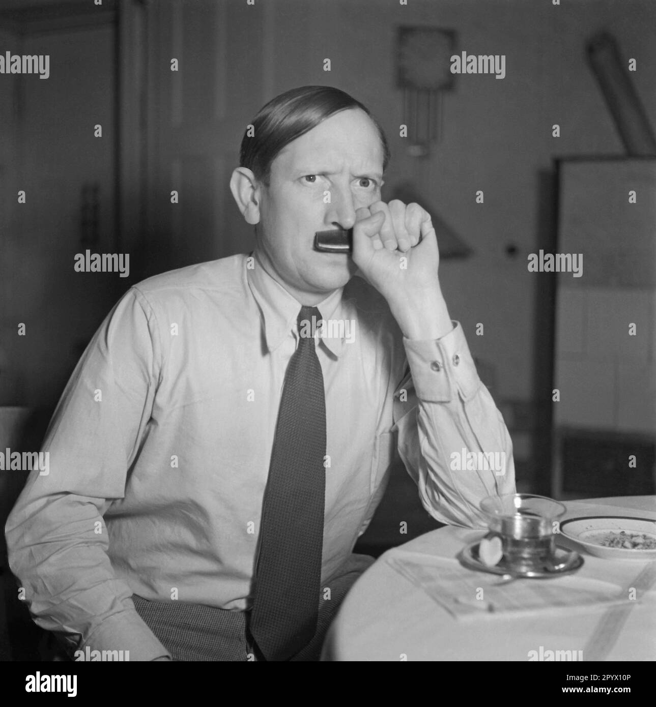Ein Mann sitzt an einem Tisch und imitiert Adolf Hitler mit seiner Frisur und einem Kamm, den er an seiner Oberlippe hält. Stockfoto