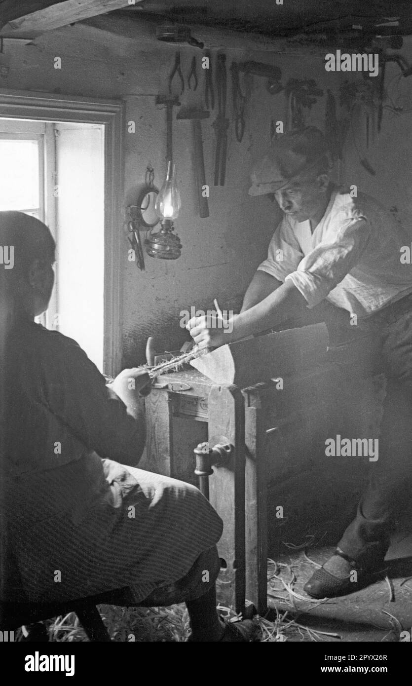 Ein Mann in schlesischem Albendorf arbeitet zusammen mit einer Frau an einem Stück Holz. Die Schnur zeigt an, dass es sich um ein Drehwerk handelt. An der Wand hängen mehrere Werkzeuge. Stockfoto