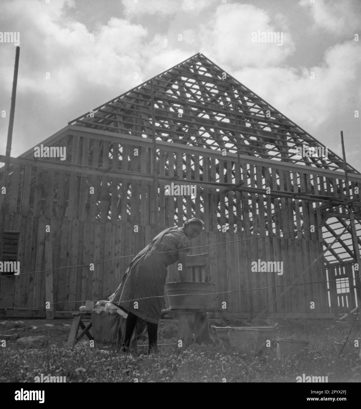 Eine Frau wäscht Kleidung vor einem Holzhaus, das noch gebaut wird. Unbezahltes Foto, wahrscheinlich 1934. [Maschinelle Übersetzung] Stockfoto