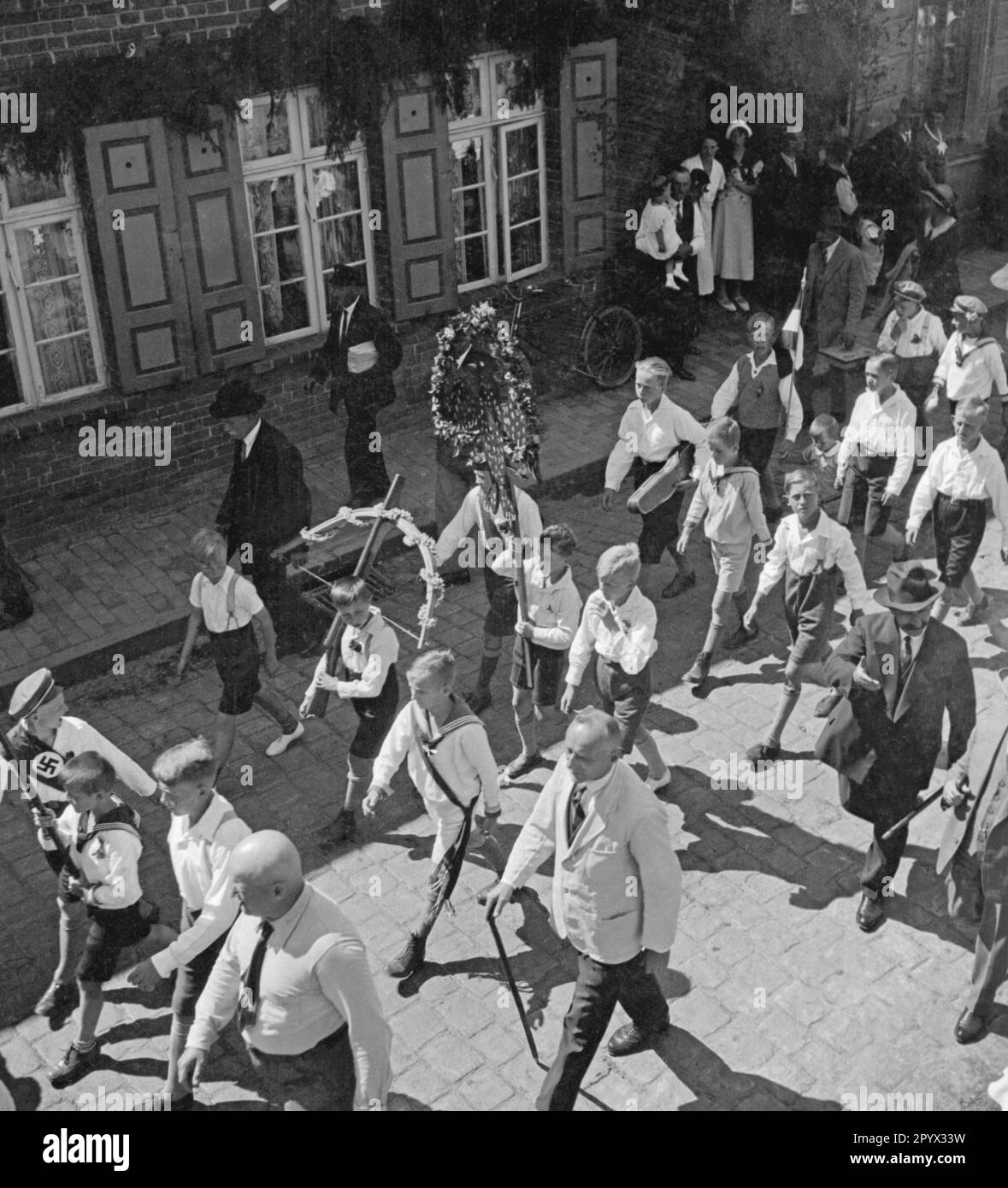 Eine Gruppe von Kindern und Jugendlichen marschiert im Rahmen des Kinderfestivals in Plau in Mecklenburg mit Blumenkränzen und Schärfe durch die Straßen. Ein Junge trägt ein Gewehr mit Schleife, auf der linken Seite ist eine Schärfe mit Hakenkreuz drauf (unbefristetes Bild). Stockfoto