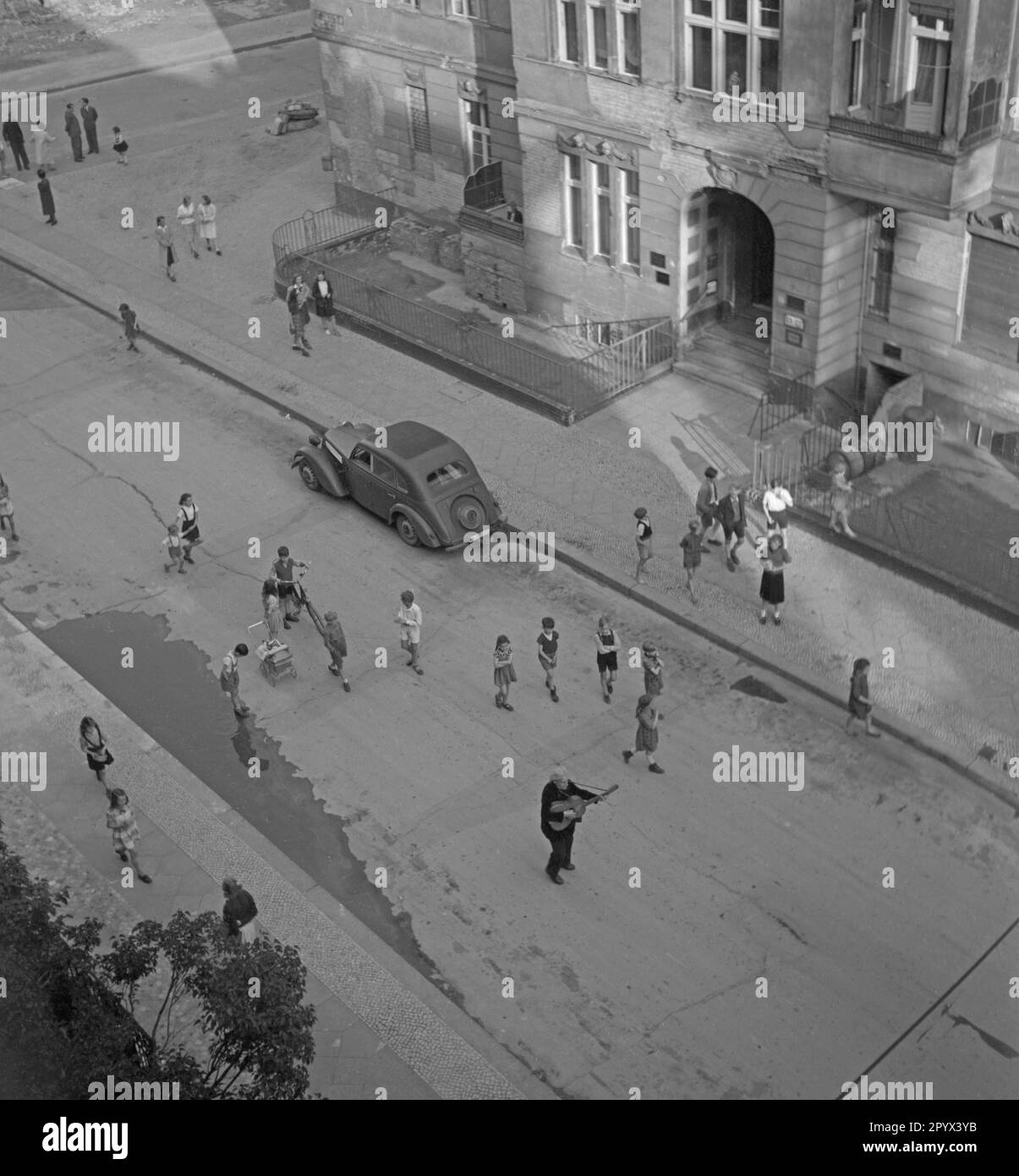 Unbezahltes Foto einer Gruppe spielender Kinder, die in einer Straße in Berlin einem Gitarrist hinterherjagen. Auf dem Bürgersteig, Passanten. Oben eine Trümmerfraktur. Stockfoto