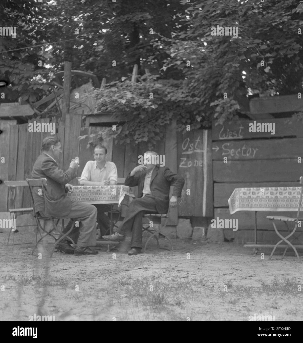 Foto einer Gruppe von Männern, die Bier am Tisch eines Restaurants für Tagesausflüge zwischen Ost- und Westberlin trinken, 1950. Die Männer sitzen an ihrem Tisch direkt an der Grenze zwischen den westlichen Sektoren und der sowjetischen Besatzungszone oder der DDR. Hinter ihnen auf den Holzplanken, die Grenze mit Kreide gezogen. Links, Osten, rechts, West. Stockfoto