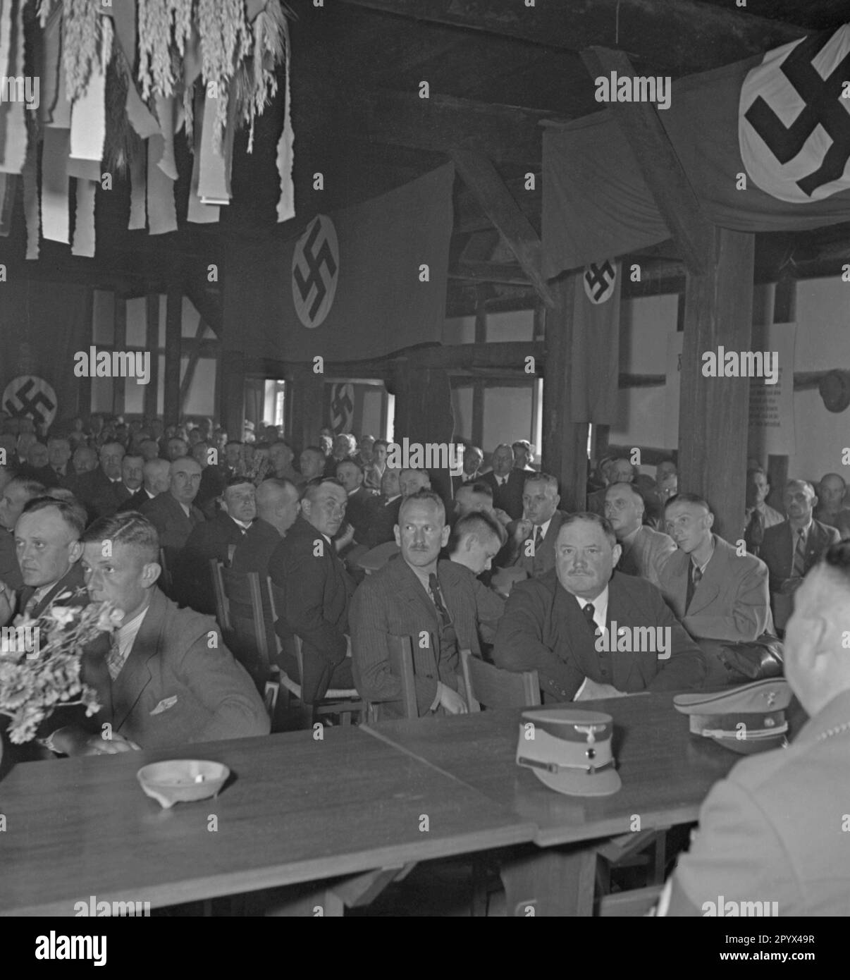 Foto eines Bauernfestivals in einer Scheune anlässlich der Eröffnung des Landtagsplatzes in Hoesseringen bei Suderburg in der Heide Lueneburg am 28. Juni 1936. An der Vorderseite sitzende Vertreter der SS und SA (Caps). Im Hintergrund Hakenkreuz-Flaggen. Stockfoto