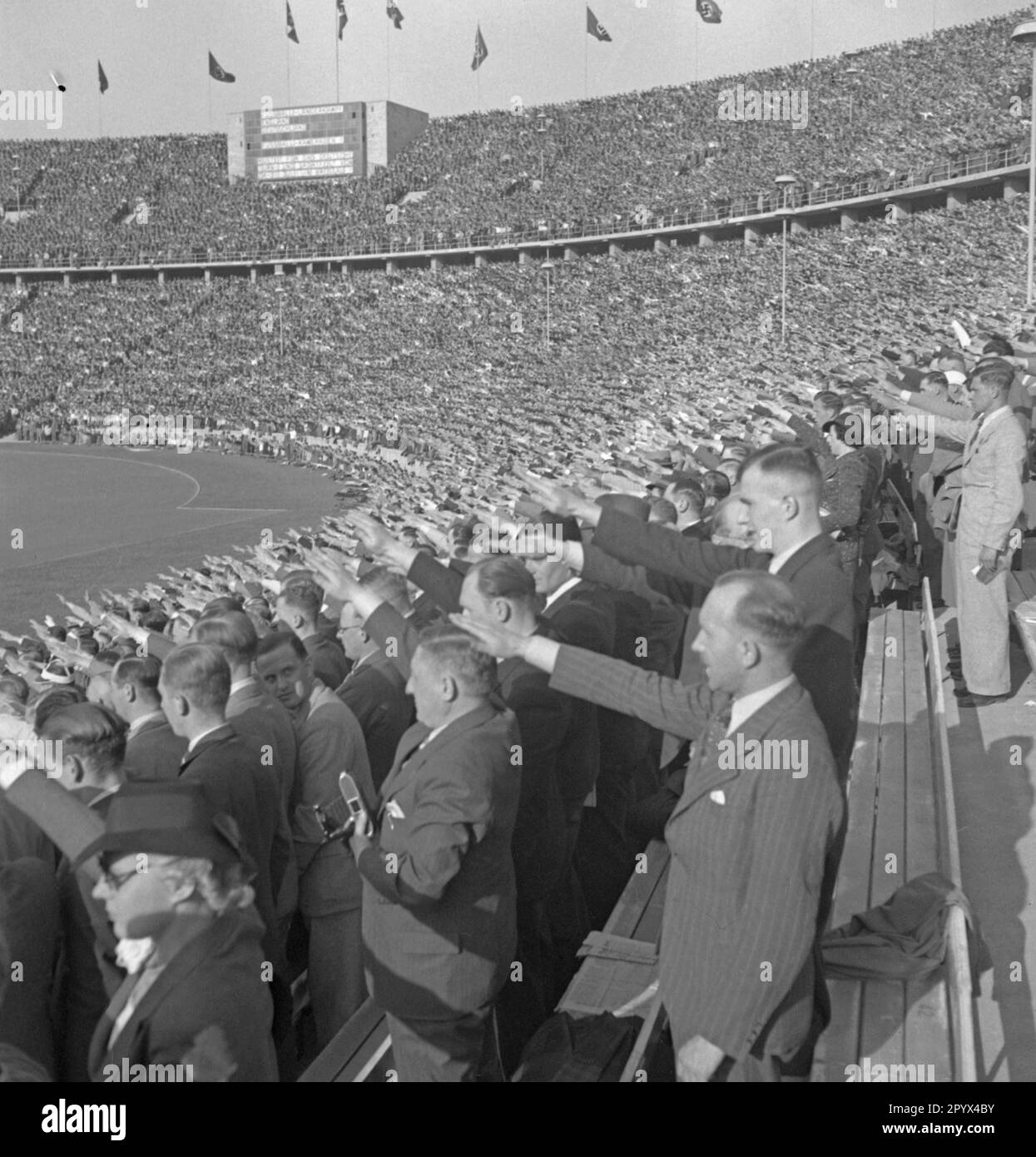 Foto der Zuschauer eines Fußballspiels im Berliner Olympiastadion (vermutlich die deutsche Nationalmannschaft) der XI Moderne Olympische Sommerspiele im August 1936. Die Zuschauer am südlichen Ende des Stadions salutieren dem Hitler. Im Hintergrund das Westende des Stadions mit der Anzeigetafel. Hakenkreuz-Flaggen wehen auf dem Gebäude. Stockfoto