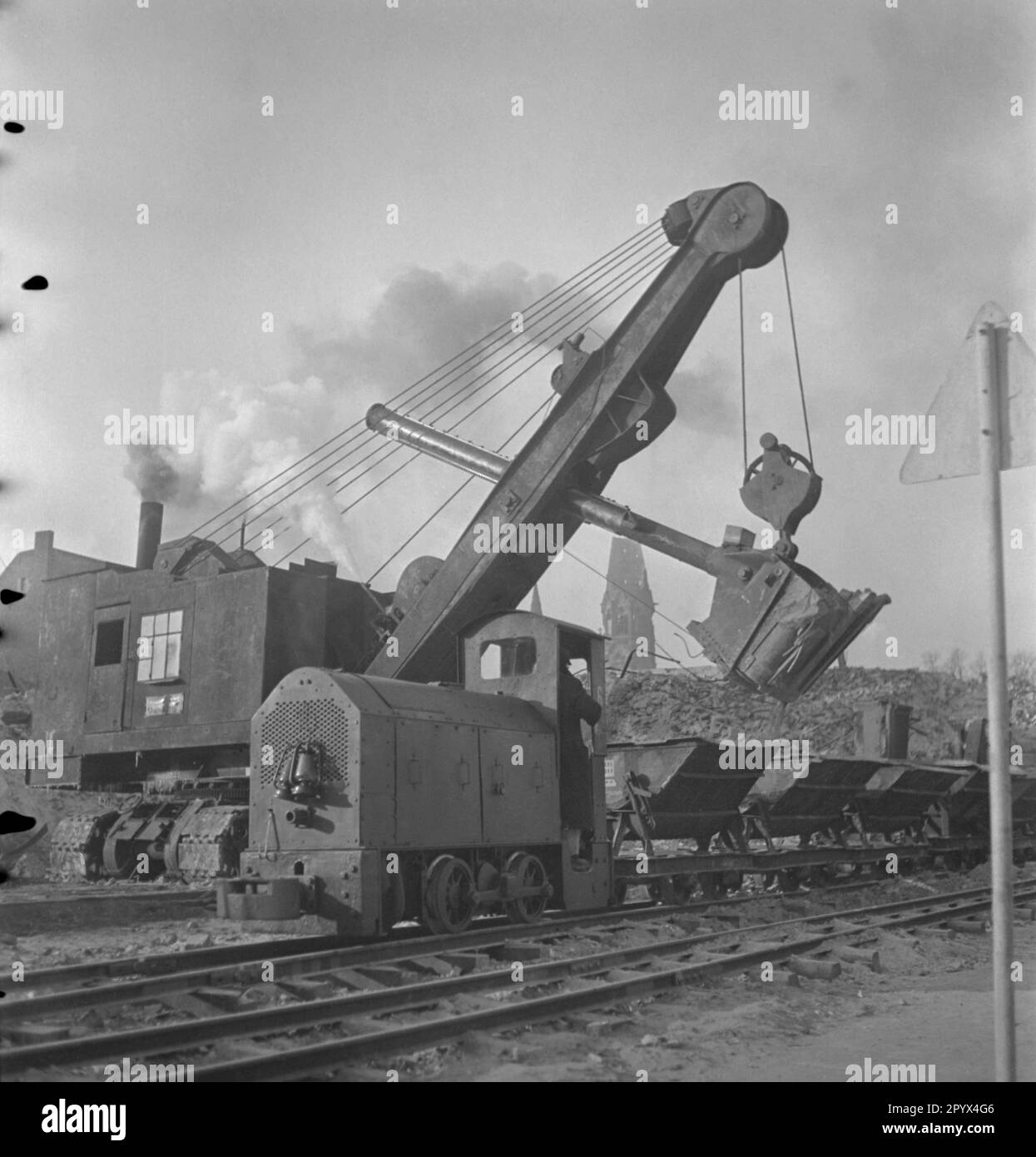 'Undatiertes Foto eines Baggers, der einen Haufen Trümmer entlädt und Wagen mit Trümmer vor einem teilweise zerstörten Wohnblock lädt, durch Bombardierung in der Nähe der Ruinen der Kaiser-Wilhelm-Gedächtniskirche am Kurfürstendamm in Charlottenburg, West-Berlin, eine ehemalige britische Besatzungszone. Die dieselbetriebene Baumaschine nimmt Ziegel und Kies mit einem Greifer auf. Im Vordergrund, Ketten und Führungsschienen. Eine schmale Diesel-Hydrauliklokomotive zieht die Wagen. Im Hintergrund befindet sich der Turm (der hohle Zahn - „Hohlzahn“) der Ruine der Kaiser-Wilhelm-Gedächtniskirche.“ Stockfoto