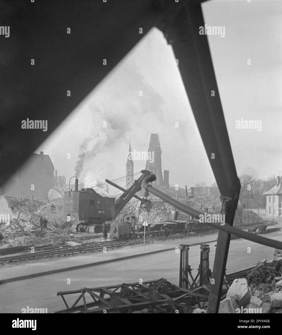 Undatiertes Foto eines Baggers, der einen Haufen Trümmer entlädt und Wagen mit Trümmer aus einem teilweise bombardierten Wohnblock in der Nähe des Kurfürstendamms im Bezirk Charlottenburg in Westberlin, einer damaligen britischen Besatzungszone, beladen. Die dieselbetriebene Baumaschine nimmt Ziegel und Kies mit einem Greifer auf. Eine schmale Diesel-Hydrauliklokomotive zieht die Wagen. Im Hintergrund befindet sich der Turm (Hohler Zahn) der Ruine der Kaiser-Wilhelm-Gedächtniskirche. Stockfoto