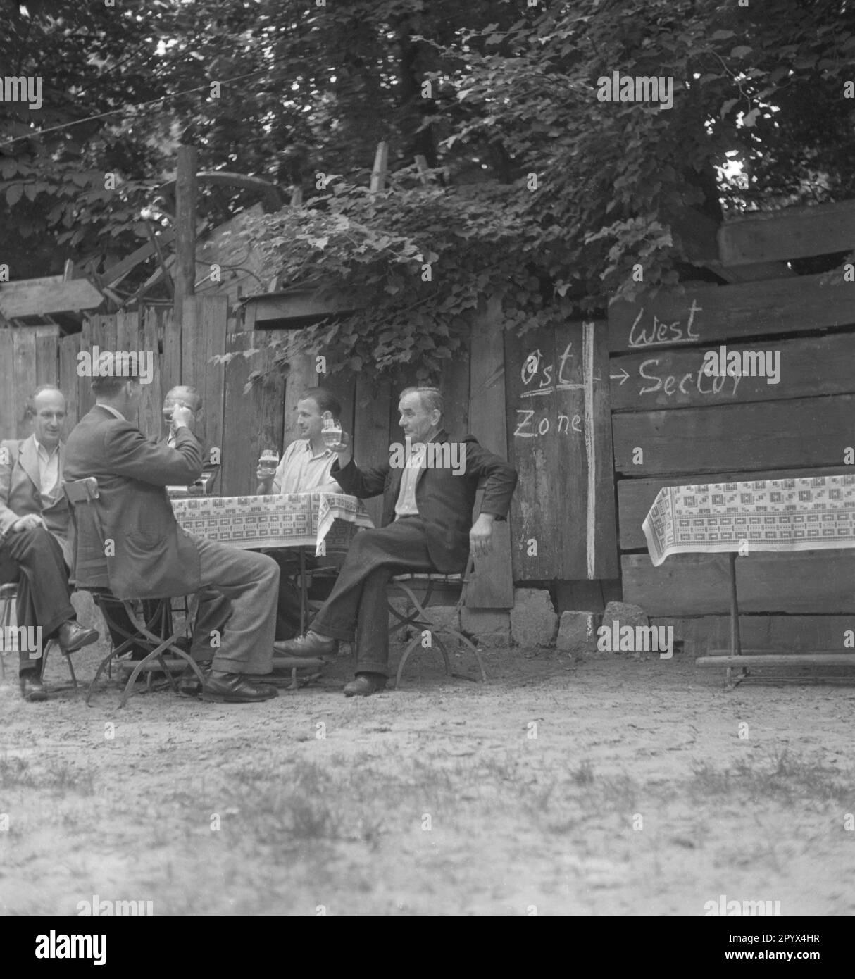 Foto einer Gruppe von Männern, die Bier am Tisch eines Restaurants für Tagesausflüge zwischen Ost- und Westberlin trinken, 1950. Die Männer sitzen an ihrem Tisch direkt an der Grenze zwischen den westlichen Sektoren und der sowjetischen Besatzungszone oder der DDR. Hinter ihnen auf den Holzplanken, die Grenze mit Kreide gezogen. Links, Osten, rechts, West. Stockfoto