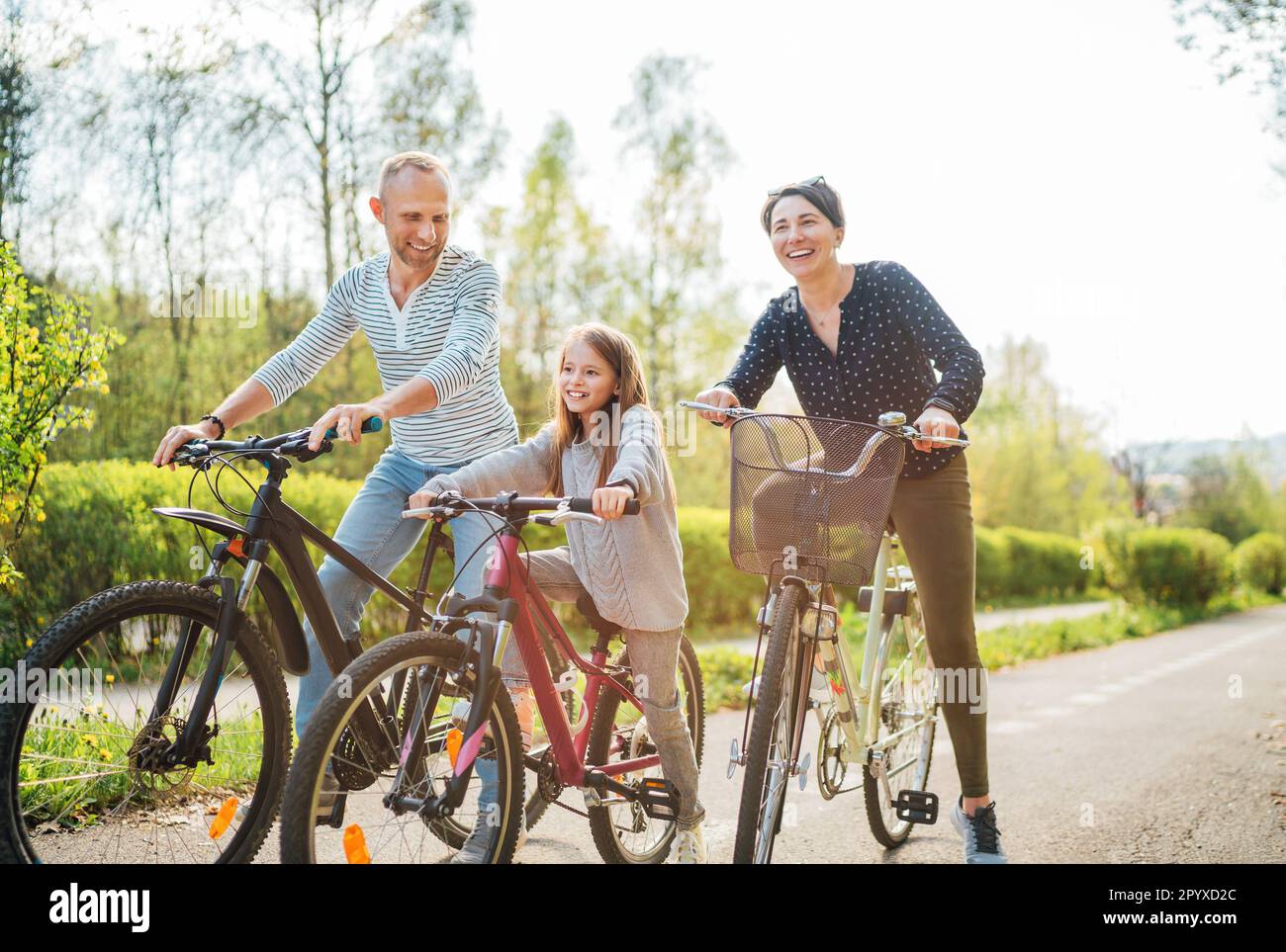 Lächelnder Vater und lächelnde Mutter mit Tochter während der Sommerfahrradrundfahrt. Sie genießen die Zusammengehörigkeit im Sommer-Stadtpark. Glückliche Elternschaft und ch Stockfoto