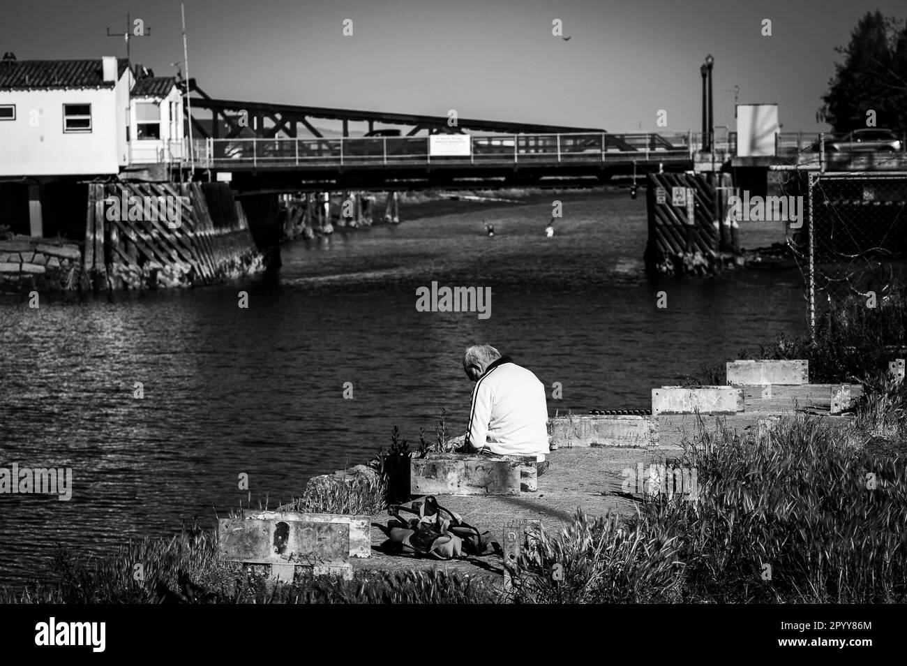 Ein erwachsener Mann in einer nachdenklichen Pose, der am Rande eines Piers mit Blick auf das ruhige Wasser eines Sees oder Meeres sitzt Stockfoto