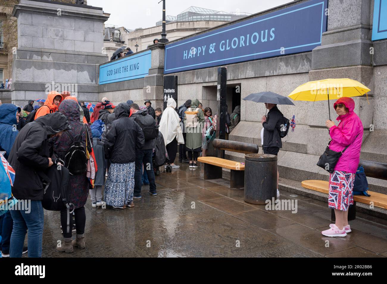 Unter dem Banner, das „glücklich und glorreich“ verkündet, stehen Mitglieder der öffentlichen Schlange für die Toiletten, nachdem sie die Krönungszeit von König Karl III. Am Trafalgar Square am 6. Mai 2023 in London, England, angeschaut haben. Stockfoto